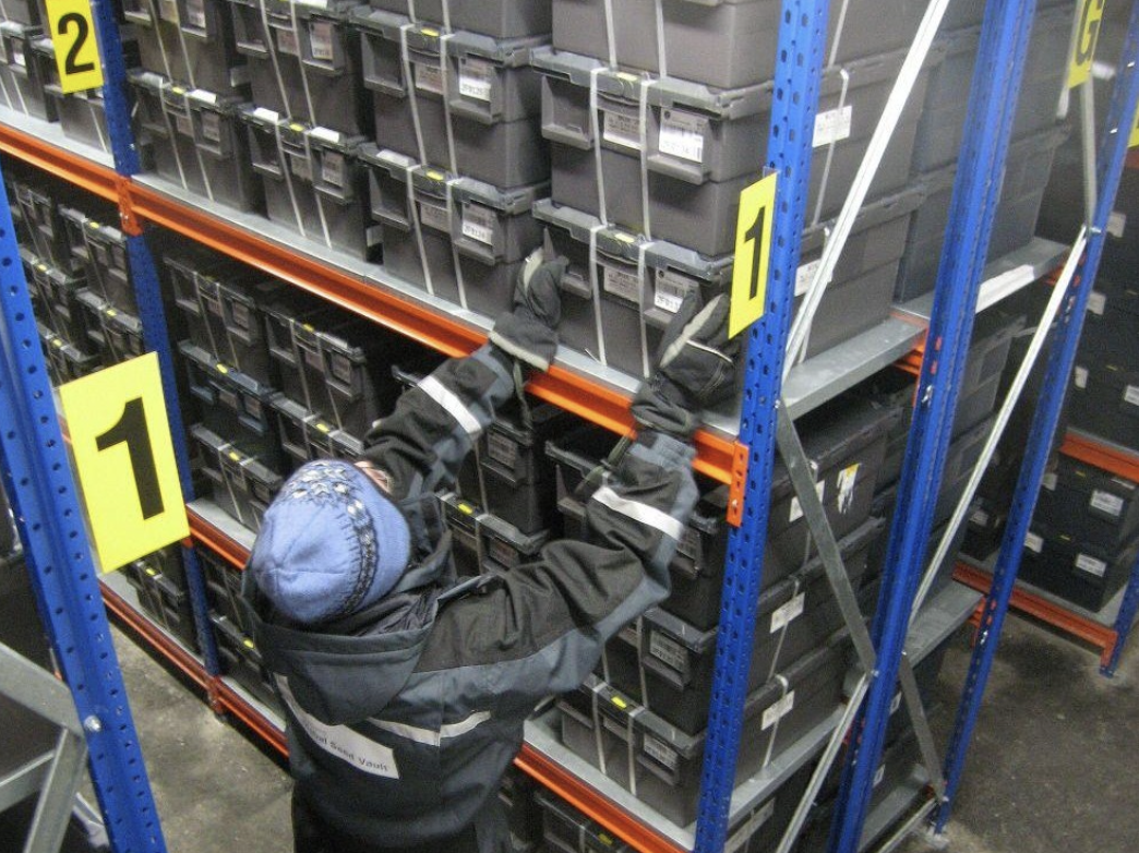 A worker in winter clothing lifts a grey storage box onto blue racking stacked with sealed seed boxes inside the Svalbard Global Seed Vault.